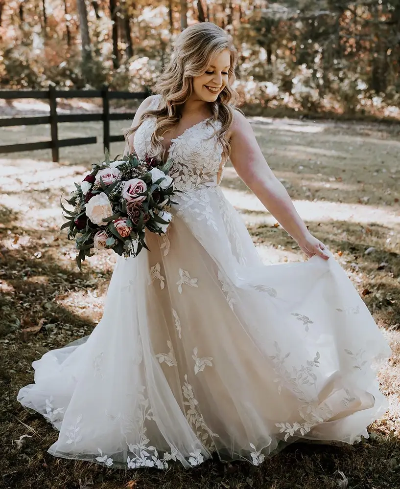 A woman in a white floral wedding dress holds a bouquet of flowers and smiles while standing outdoors on a grassy area with trees and a wooden fence in the background. - Chevalrie Salon and Spa