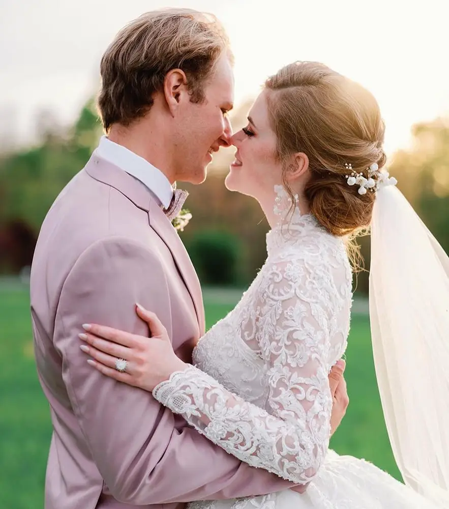 A bride and groom in formal wedding attire embrace outdoors, touching noses and smiling, with sunlight in the background. - Chevalrie Salon and Spa