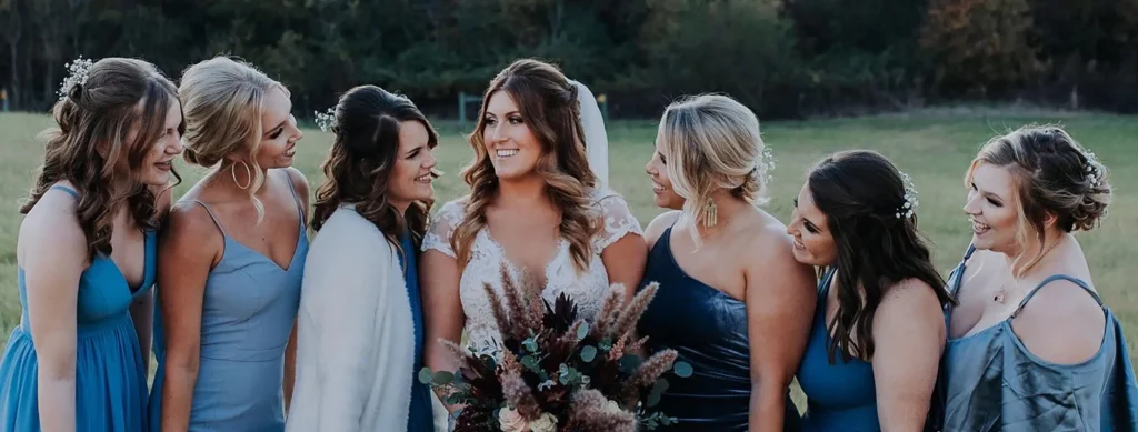 A bride in a white dress holds a bouquet and stands outdoors, surrounded by six women in blue dresses, all smiling and looking at her. - Chevalrie Salon and Spa
