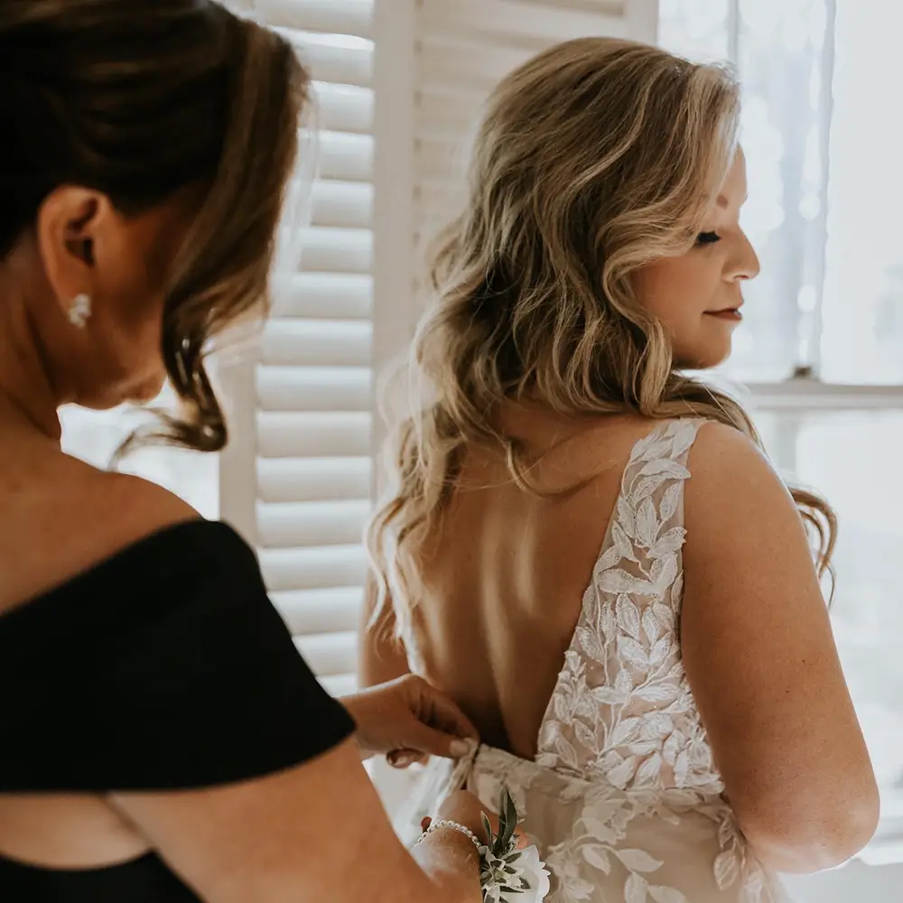 A woman in a white lace dress stands as another woman fastens the back of her dress near a window with white shutters. - Chevalrie Salon and Spa