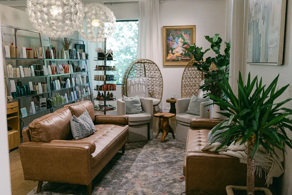 A cozy salon waiting area with two brown leather sofas, wicker chairs, shelves with hair products, potted plants, and wall art. - Chevalrie Salon and Spa