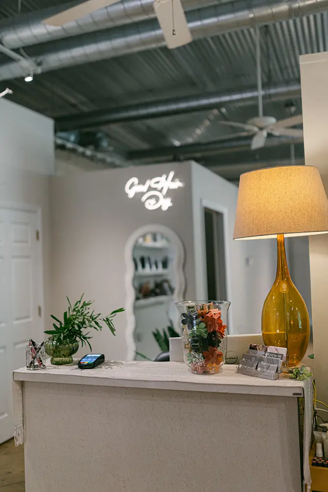 Reception desk with a lamp, plants, business cards, and a glass vase of flowers; a sign and mirror are visible on the back wall in a modern interior space. - Chevalrie Salon and Spa