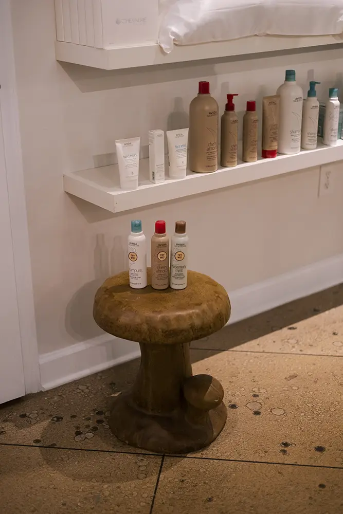 Assorted hair care products are displayed on two shelves and a small mushroom-shaped stool against a wall in a tiled room. - Chevalrie Salon and Spa