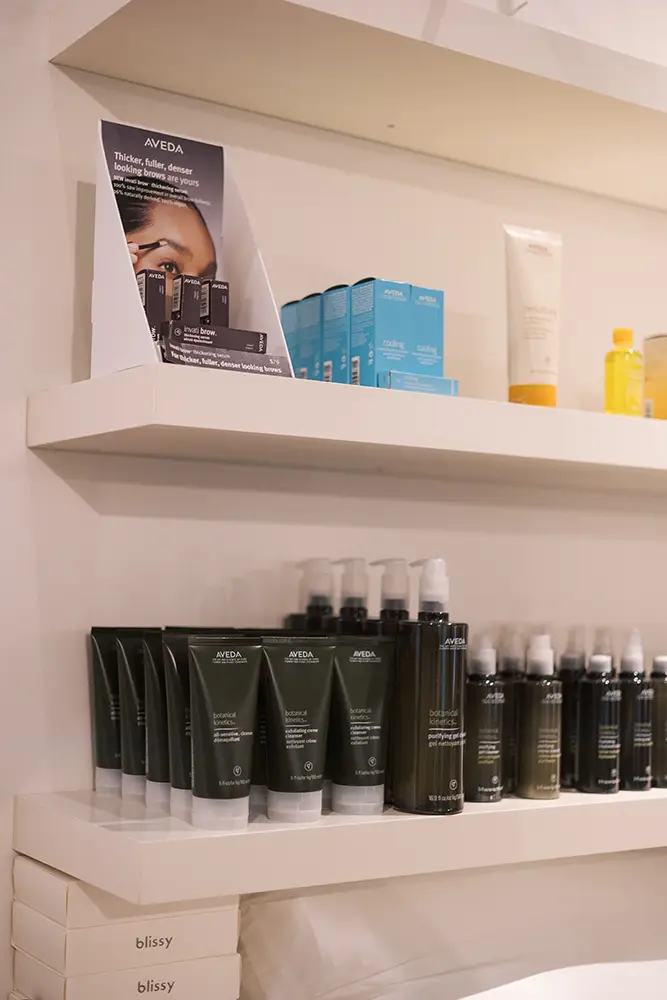 Shelves display various beauty and haircare products, including Aveda bottles, blue boxes, and other skincare items, arranged neatly in a retail setting. - Chevalrie Salon and Spa