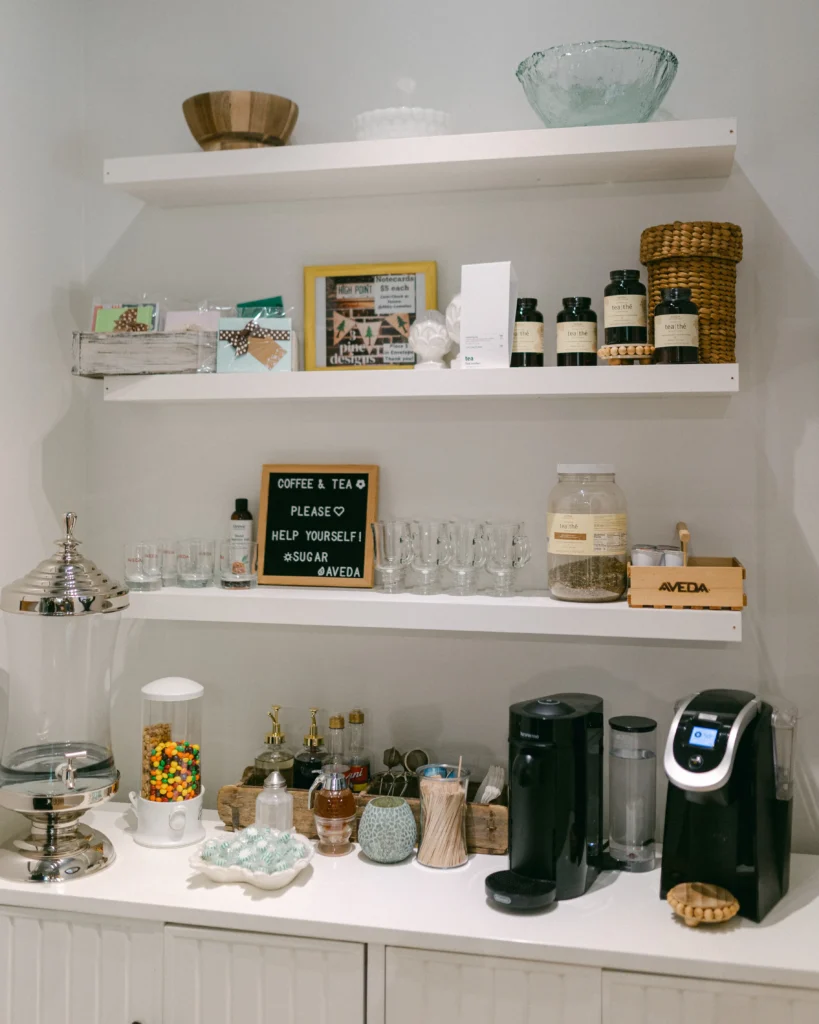 A coffee and tea station with white shelves holding jars, cups, and tea supplies, plus coffee machines, a candy dispenser, and a sign inviting guests to help themselves. - Chevalrie Salon and Spa