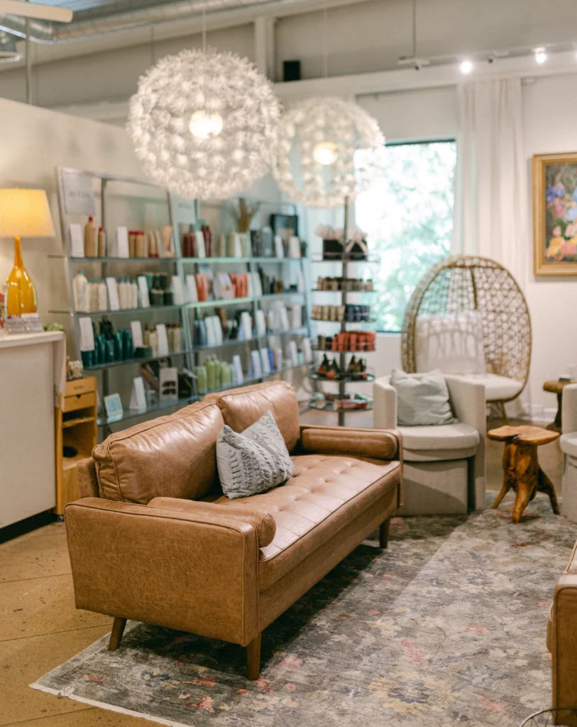 A modern salon interior with a brown leather sofa, shelves of hair products, two round pendant lights, and a patterned rug. - Chevalrie Salon and Spa