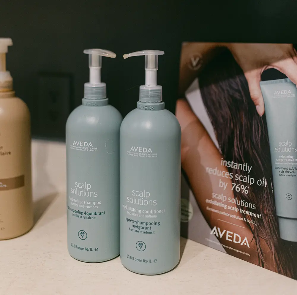 Two bottles of Aveda Scalp Solutions shampoo and conditioner with pump dispensers are displayed on a counter next to a product advertisement. - Chevalrie Salon and Spa
