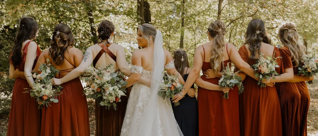 A bride and her bridesmaids stand outdoors with their backs to the camera, arms linked, holding bouquets and wearing dresses in warm earth tones. - Chevalrie Salon and Spa
