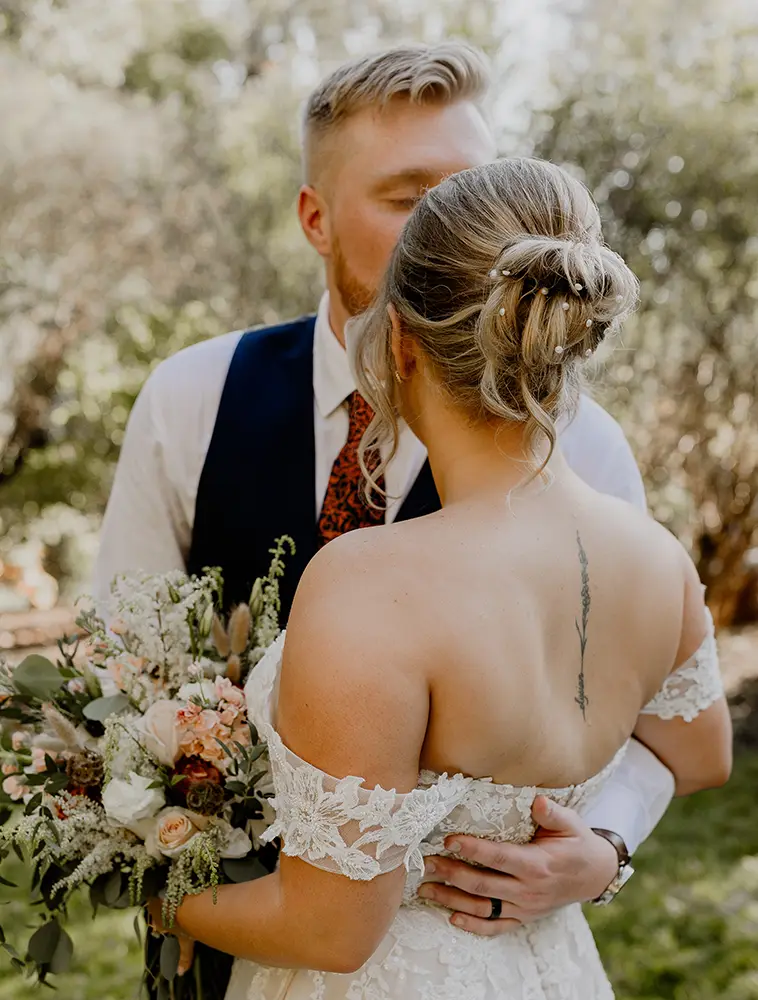 A groom kisses a bride on the head as they embrace outdoors. The bride holds a bouquet and has a floral tattoo on her upper back. - Chevalrie Salon and Spa