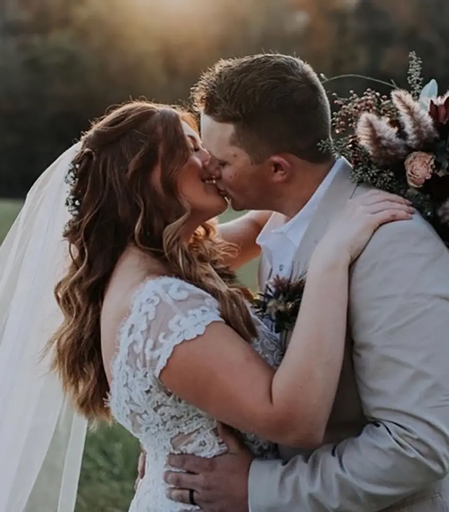 A bride and groom kiss outdoors at sunset, the bride wearing a lace dress and veil, the groom in a light suit, holding a bouquet of flowers. - Chevalrie Salon and Spa