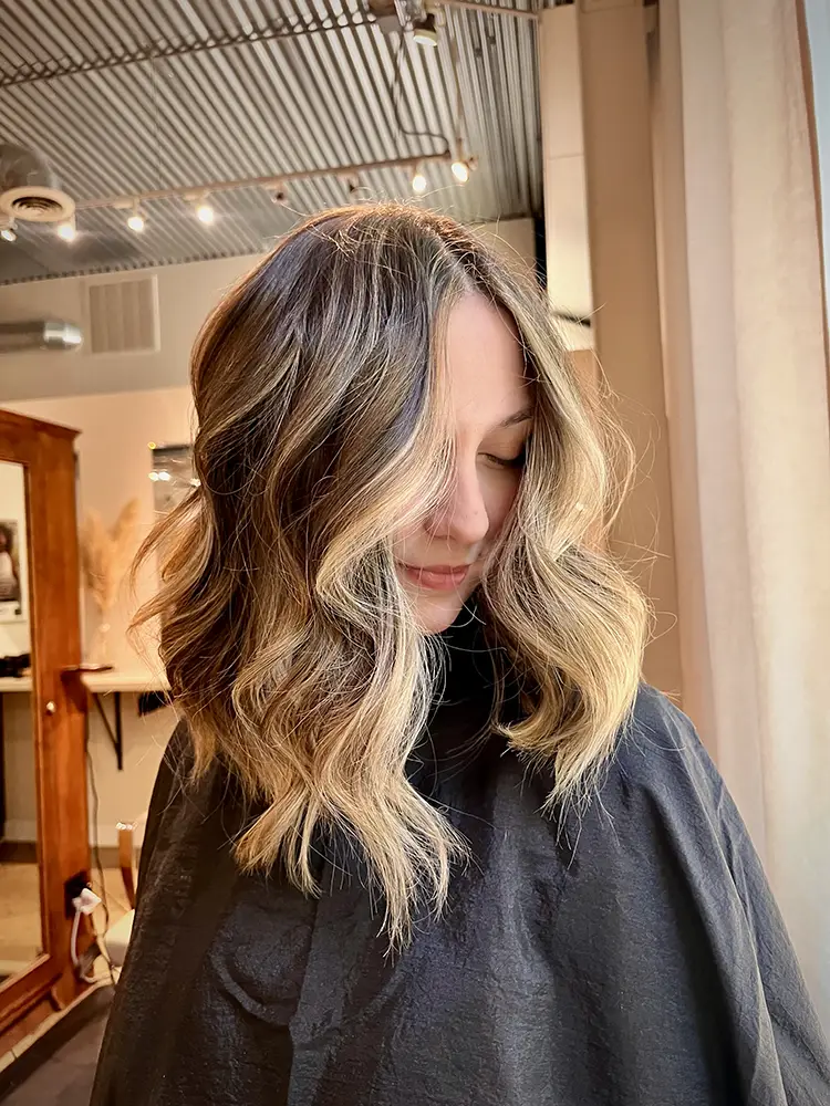 A woman with wavy, shoulder-length blonde balayage hair sits in a salon, wearing a black cape and facing slightly downward. - Chevalrie Salon and Spa