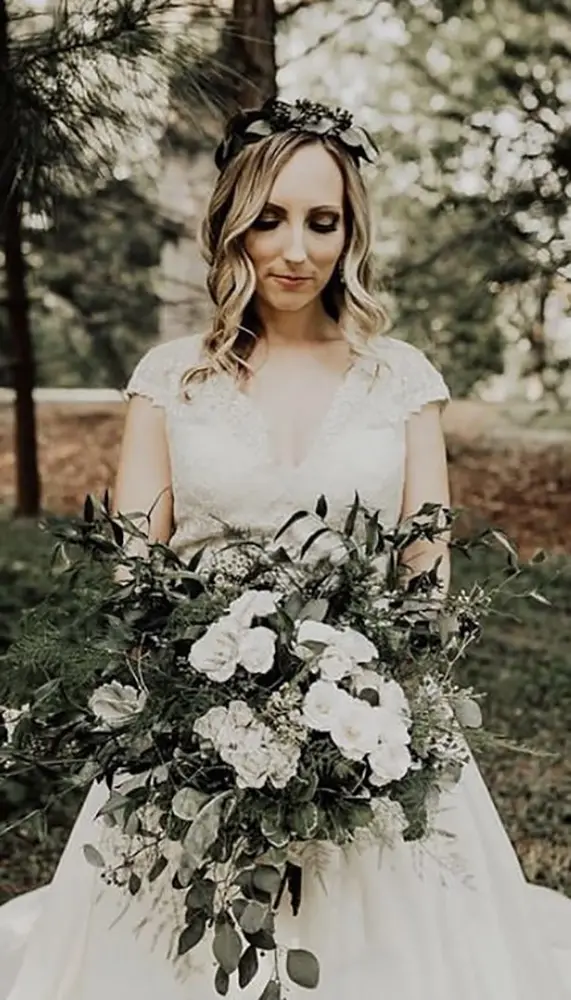 A bride in a white dress with lace details holds a large bouquet of white flowers and greenery, standing outdoors with trees in the background. - Chevalrie Salon and Spa