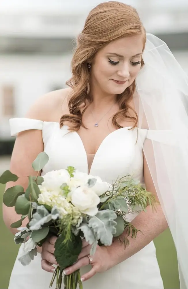A bride with red hair in an off-shoulder white gown holds a bouquet of white flowers and greenery, looking down and smiling slightly. - Chevalrie Salon and Spa