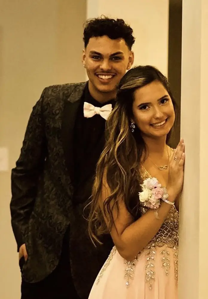 A young man in a suit and bow tie stands behind a young woman in a beaded dress with a corsage, both smiling indoors. - Chevalrie Salon and Spa