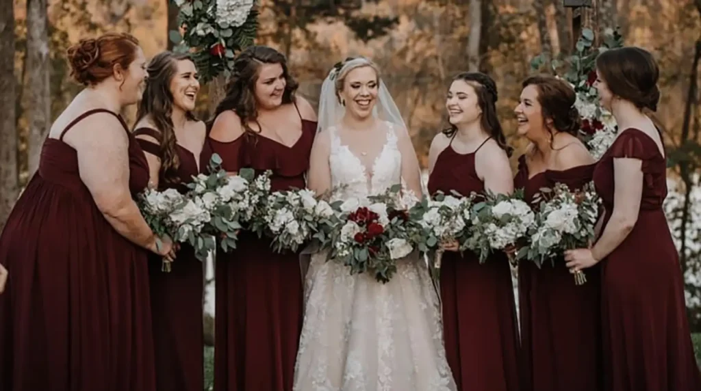 A bride in a white gown stands outdoors with six bridesmaids in matching burgundy dresses, all holding bouquets and smiling. - Chevalrie Salon and Spa