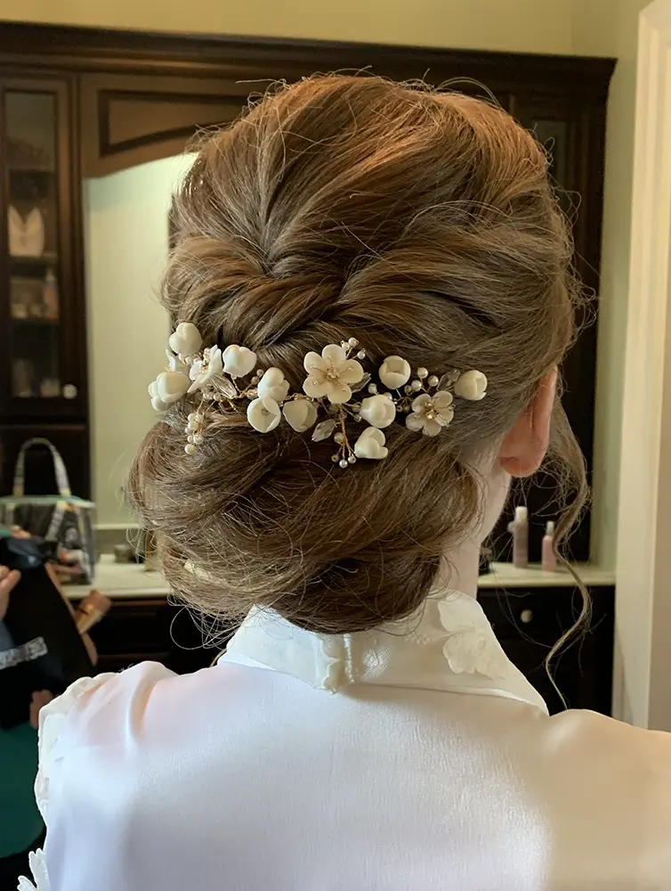 A woman with light brown hair styled in an elegant updo adorned with white floral hairpins, wearing a white robe, is seen from behind in a room with dark cabinetry. - Chevalrie Salon and Spa