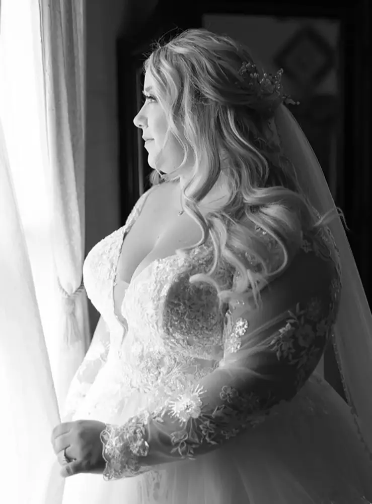 A woman in a long-sleeved lace wedding dress looks out of a window, with light illuminating her face and hair. - Chevalrie Salon and Spa