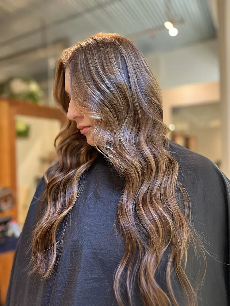 A woman with long, wavy brown hair with blonde highlights sits in a salon chair, wearing a black cape. - Chevalrie Salon and Spa