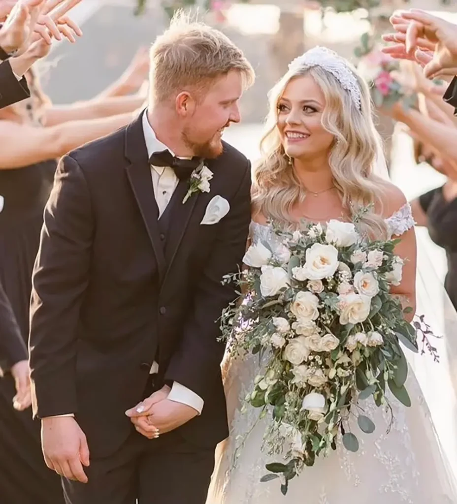 A bride and groom in formal attire smile at each other, holding hands. The bride holds a large bouquet of white flowers, and people in black outfits celebrate around them. - Chevalrie Salon and Spa