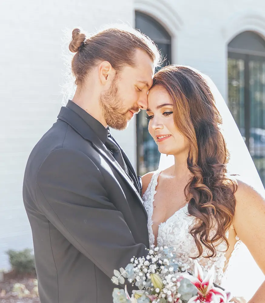 A bride and groom stand close together outdoors, with the groom in a dark suit and the bride in a white dress holding a bouquet. - Chevalrie Salon and Spa